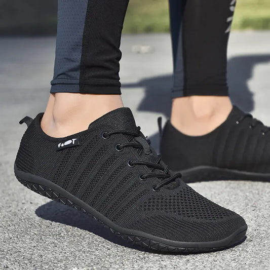 zapatillas negras mujer Barefoot de malla, ligeras y con suela plana, sobre pavimento gris.